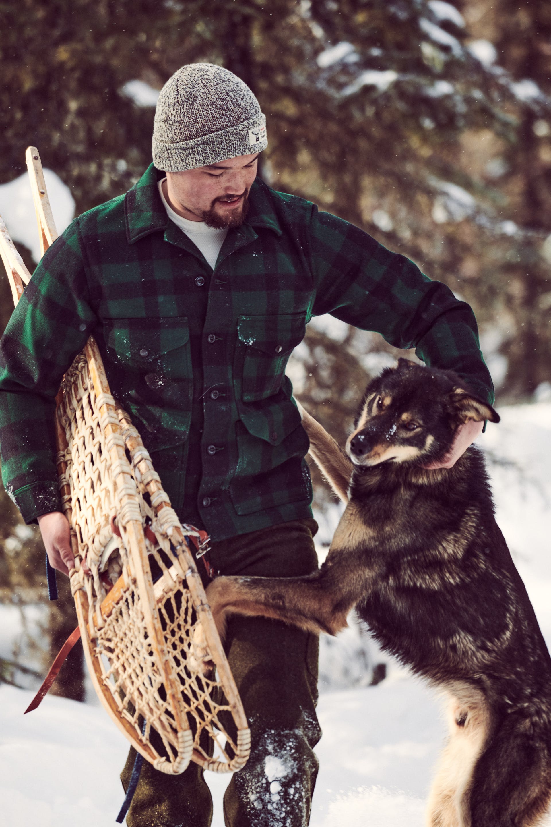 Man wearing Filson Mackinaw Wool Cruiser Jacket in green/black plaid holding snowshoes while playing with his dog