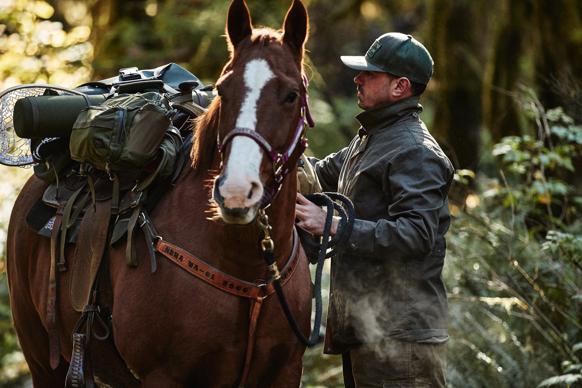 Man wearing Filson Cover Cloth Mile Marker Coat in otter green inspecting items on horse saddle